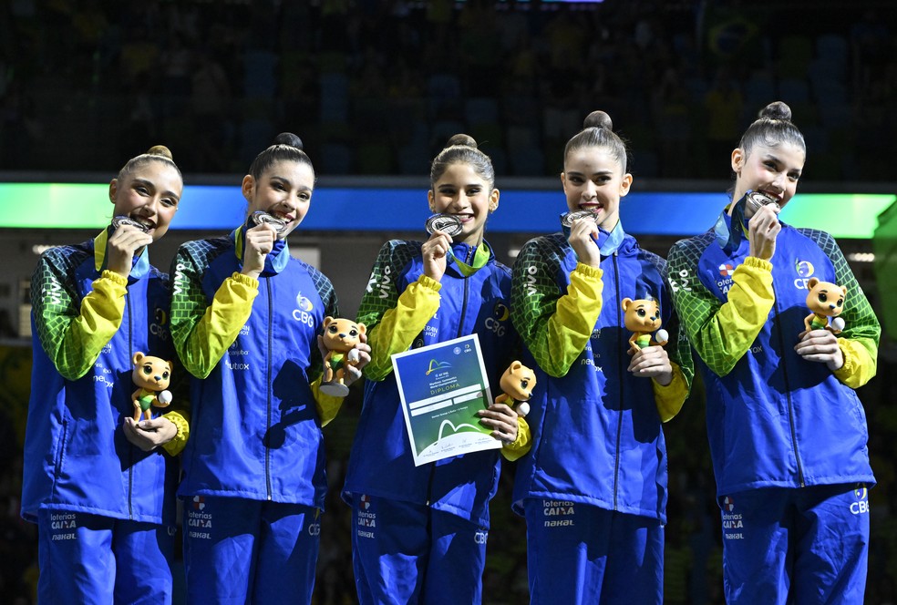 Seleção brasileira feminina de ginástica rítmica comemorando medalha histórica no Mundial 2025, no Rio de Janeiro, com torcida ao fundo.