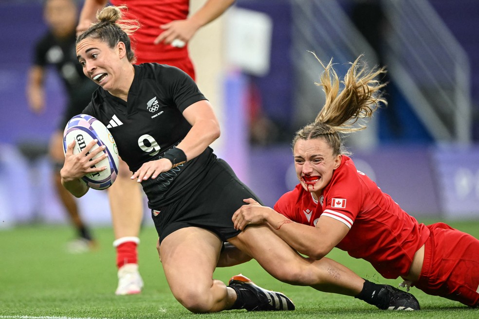 Com sangue escorrendo, canadense Krissy Scurfield tenta parar jogadora da Nova Zelândia, no duelo de rugby das Olimpíadas — Foto: Carl de Souza/AFP